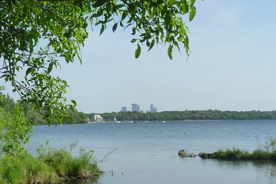 The shore of Lake Harriet with trees in the foreground and the downtown Minneapolis skyline far in the distance