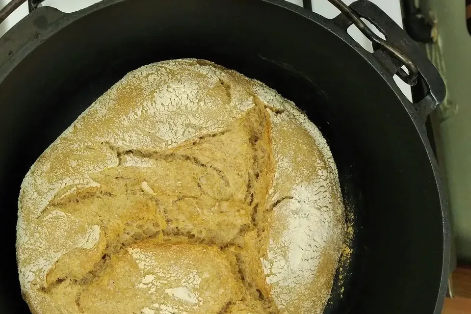 A round, scored loaf of sourdough bread with a floury golden crust sitting in a black cast-iron pot
